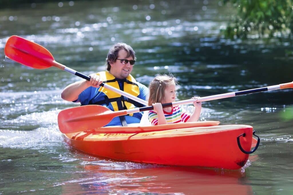 An adult and a child wearing life jackets paddle a red kayak on a calm river.