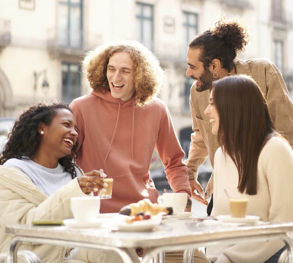 Four people enjoy drinks at an outdoor cafe, laughing and chatting together around a table with coffee cups and a plate of dessert.