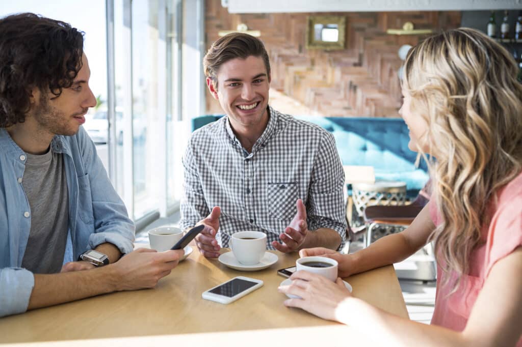 Three people sit at a cafe table, smiling and talking, with coffee cups and smartphones in front of them.