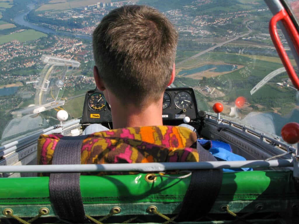 Person in a glider cockpit, flying over a landscape with rivers and houses visible below.