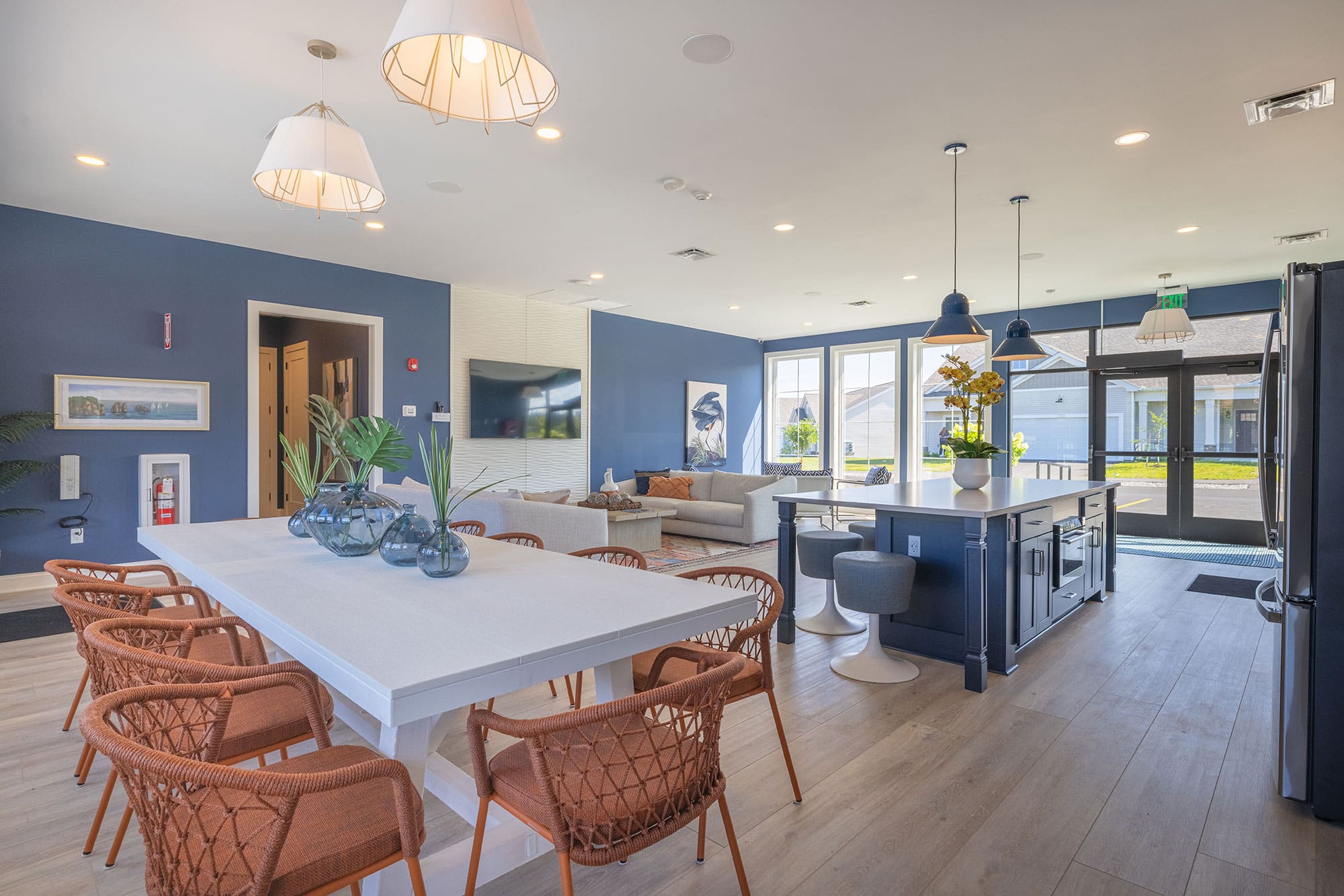 Modern open-concept room with a dining table, chairs, kitchen island, and living area featuring large windows. Blue and white decor with hanging lights.