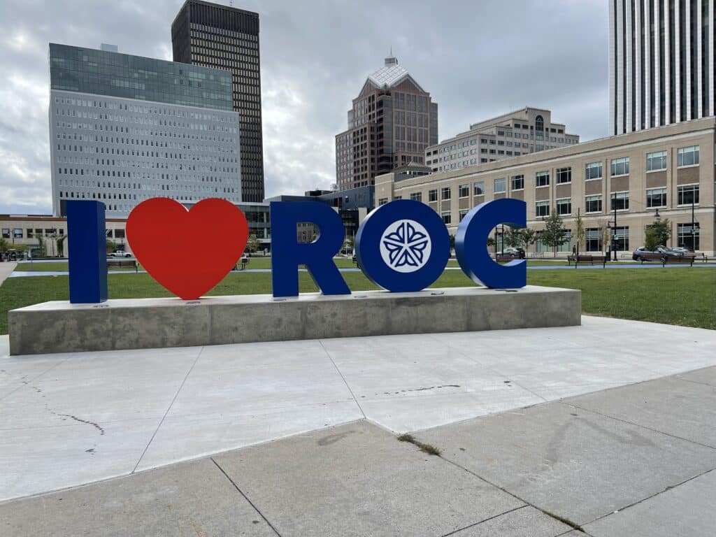 I heart ROC" sign with city buildings in the background under a cloudy sky.