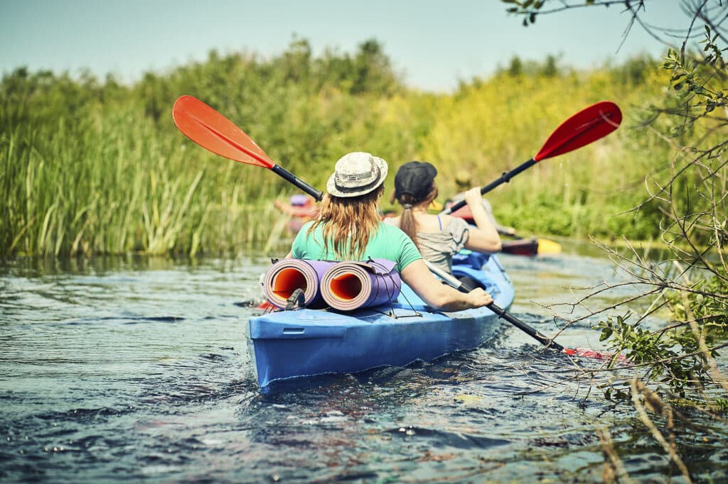 Two people kayaking on a river with backpacks and rolled mats on their kayak, surrounded by greenery.