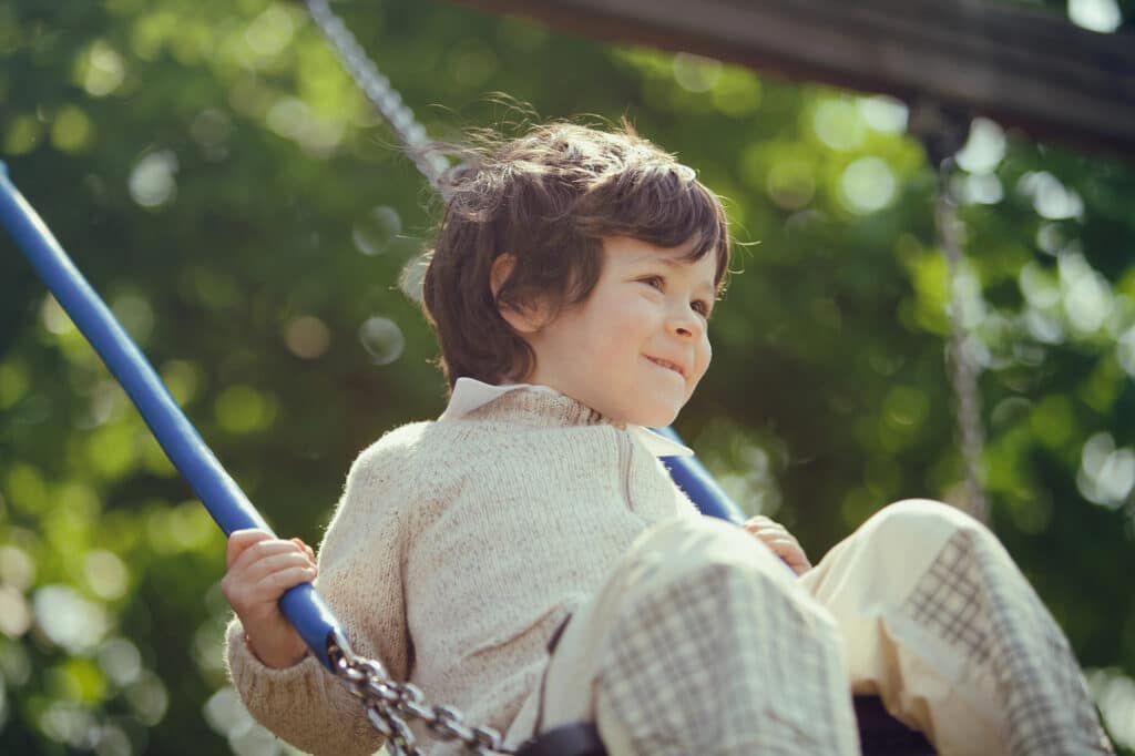 Child on a swing, smiling and holding onto blue ropes, in a sunlit park with green foliage in the background.