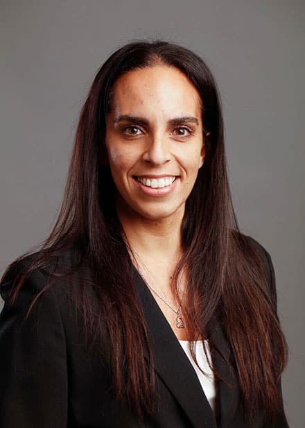 A person with long dark hair wearing a black blazer and white top smiles against a gray background.