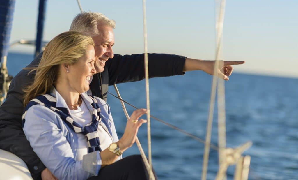 A man and woman smile and look out over the water while on a sailboat. The man points towards something in the distance.