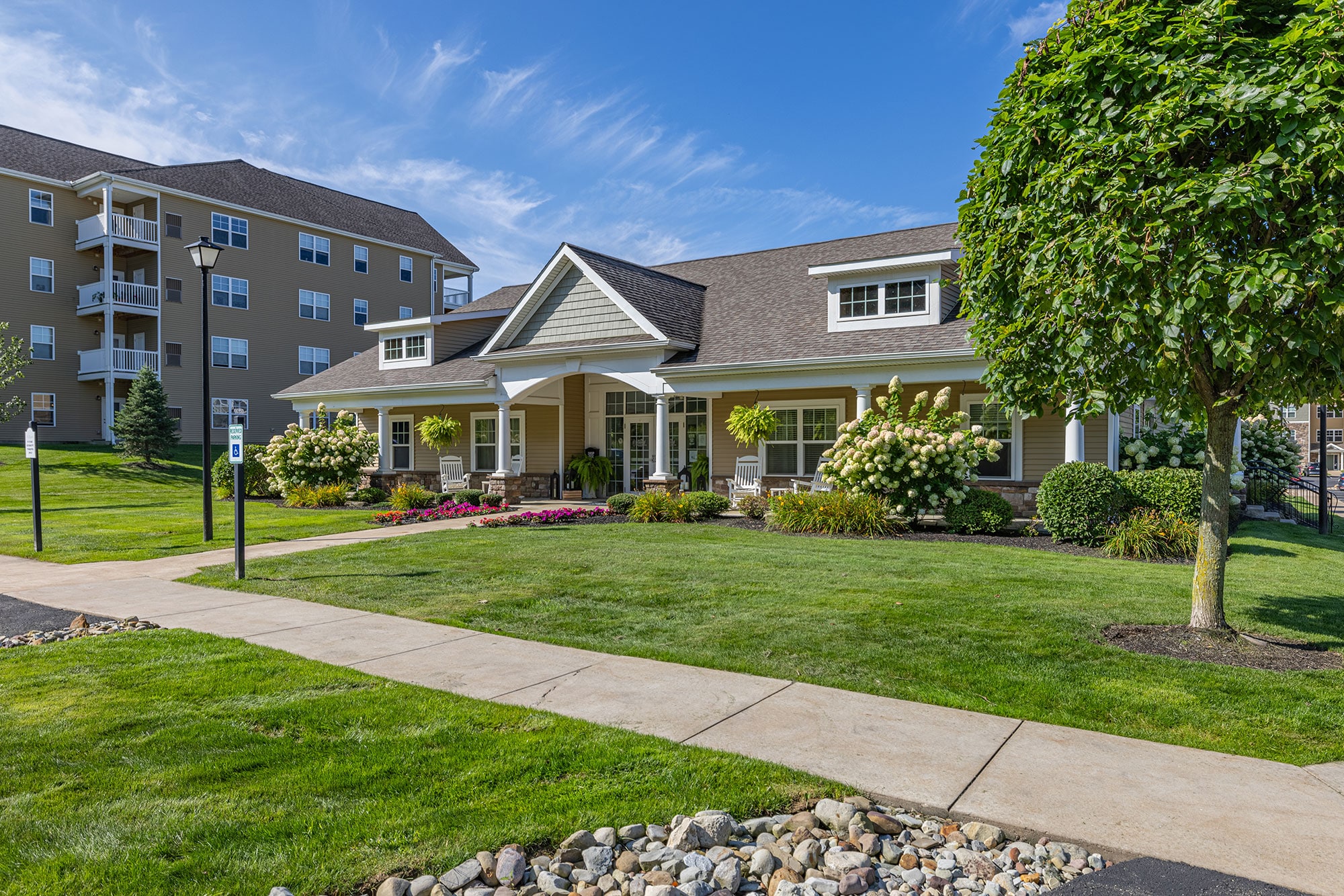 A modern suburban house with a manicured lawn, flowerbeds, and trees, set against a blue sky. An adjacent apartment building is visible in the background.