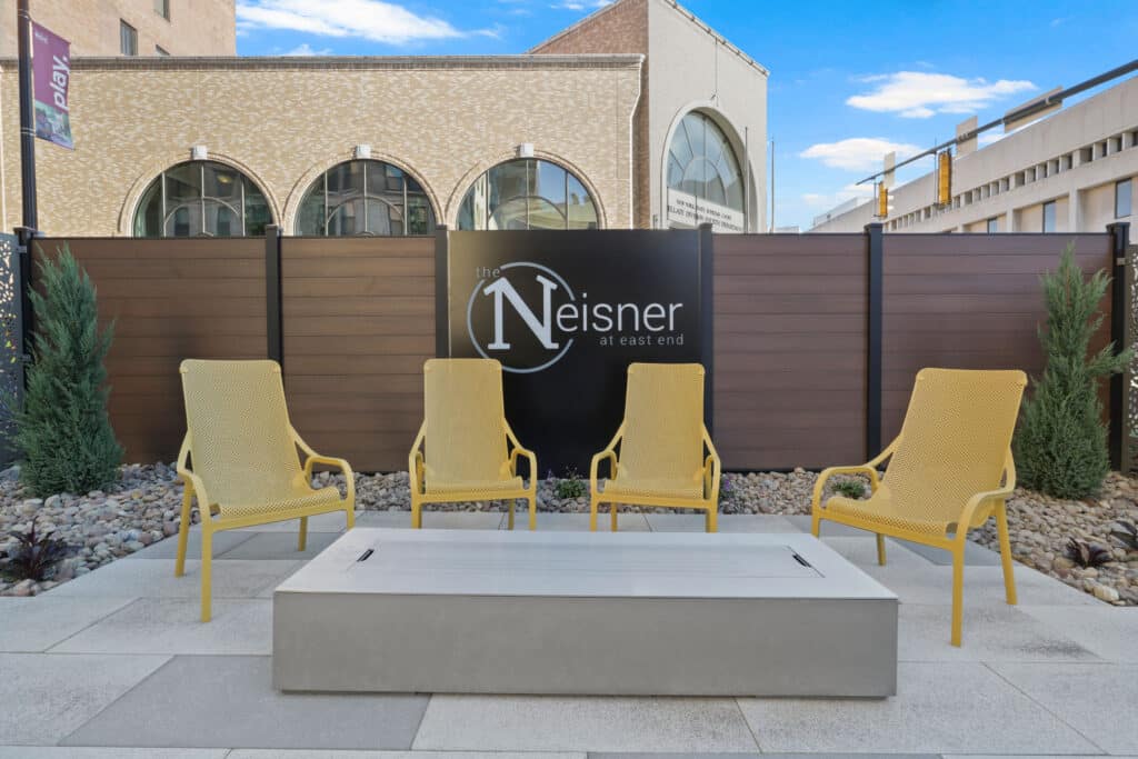 Four yellow chairs surround a rectangular concrete table in an outdoor patio area, with a sign reading "The Neisner at East End" in the background.