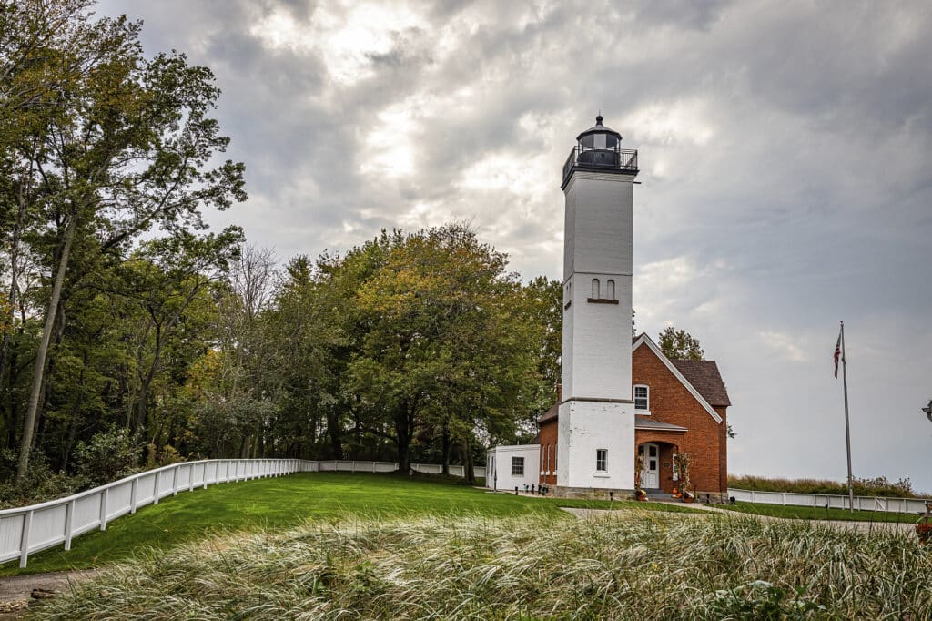 A white lighthouse with an attached brick building stands near a grassy path, surrounded by trees and a white fence under a cloudy sky.