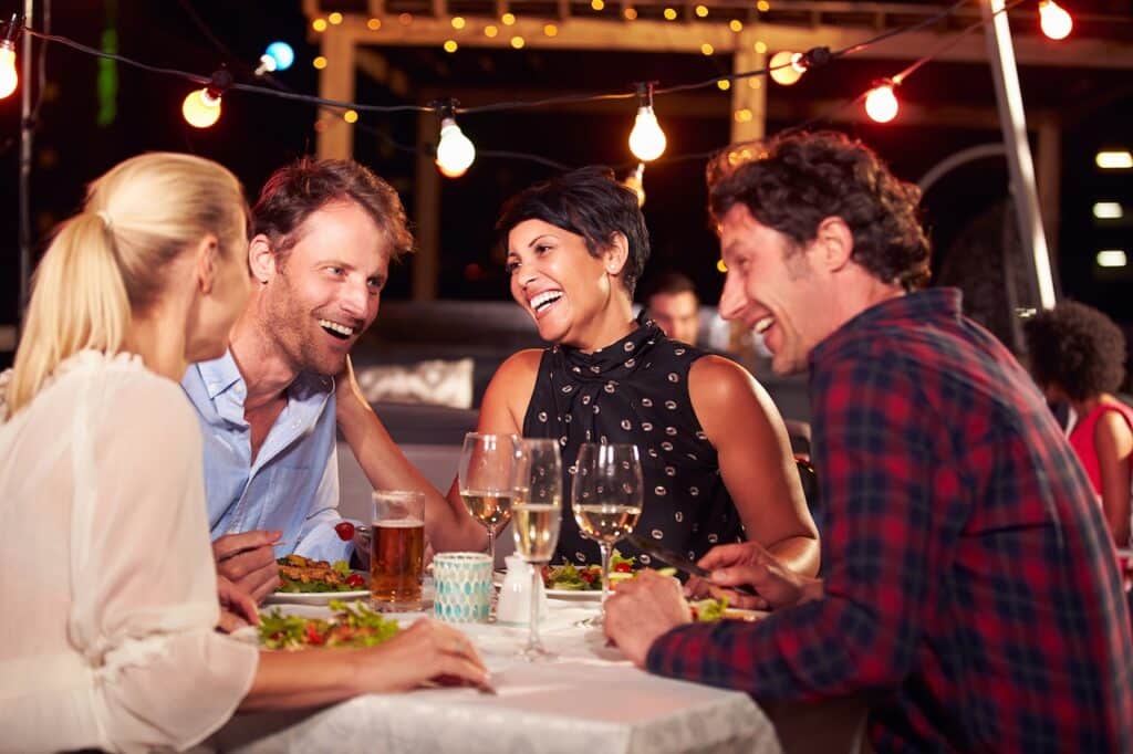 Four people enjoying dinner at an outdoor restaurant with drinks and salads on the table, surrounded by string lights.