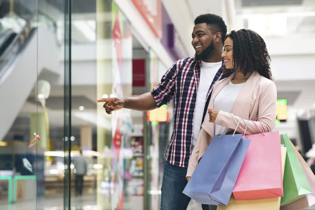 A couple stands in a shopping mall, smiling and looking at a store display. The man points excitedly, while the woman holds several shopping bags.