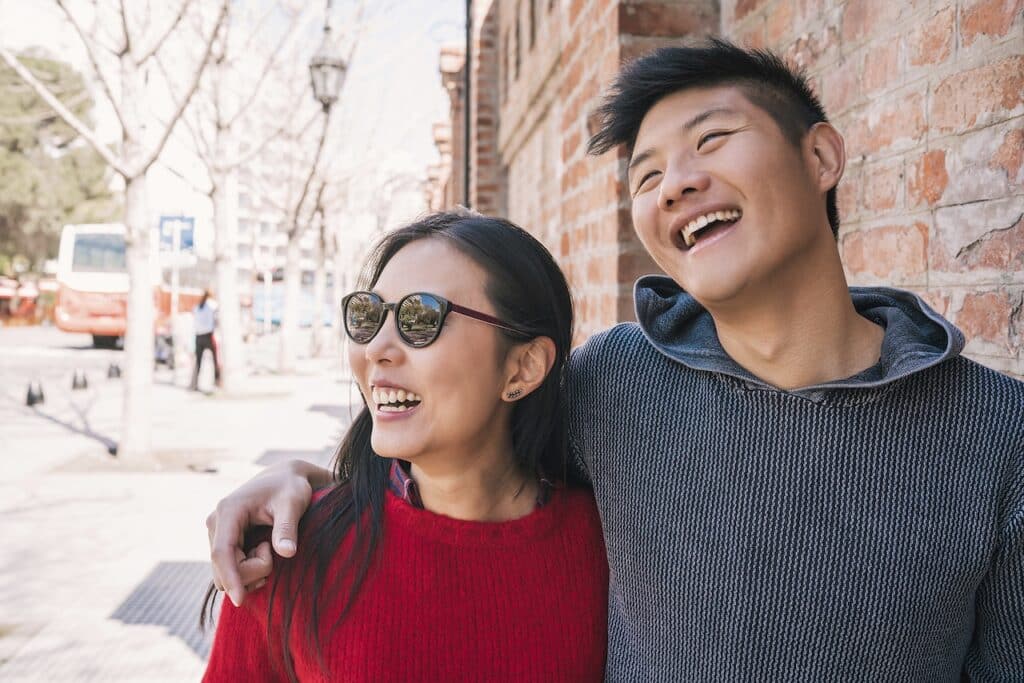 Two people smiling, standing side by side. The person on the left is in a red sweater and sunglasses, while the person on the right wears a gray sweater. They are outdoors near a brick wall.