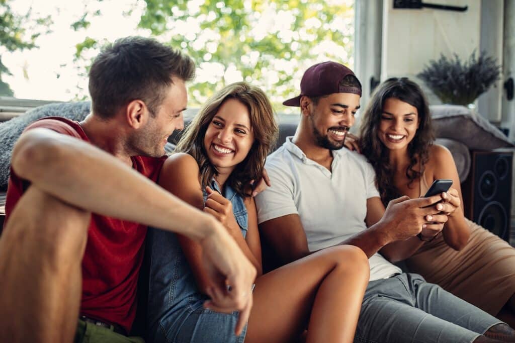 Four friends sit together on a sofa, two men and two women. One man shows something on his phone to the others, and they all smile.