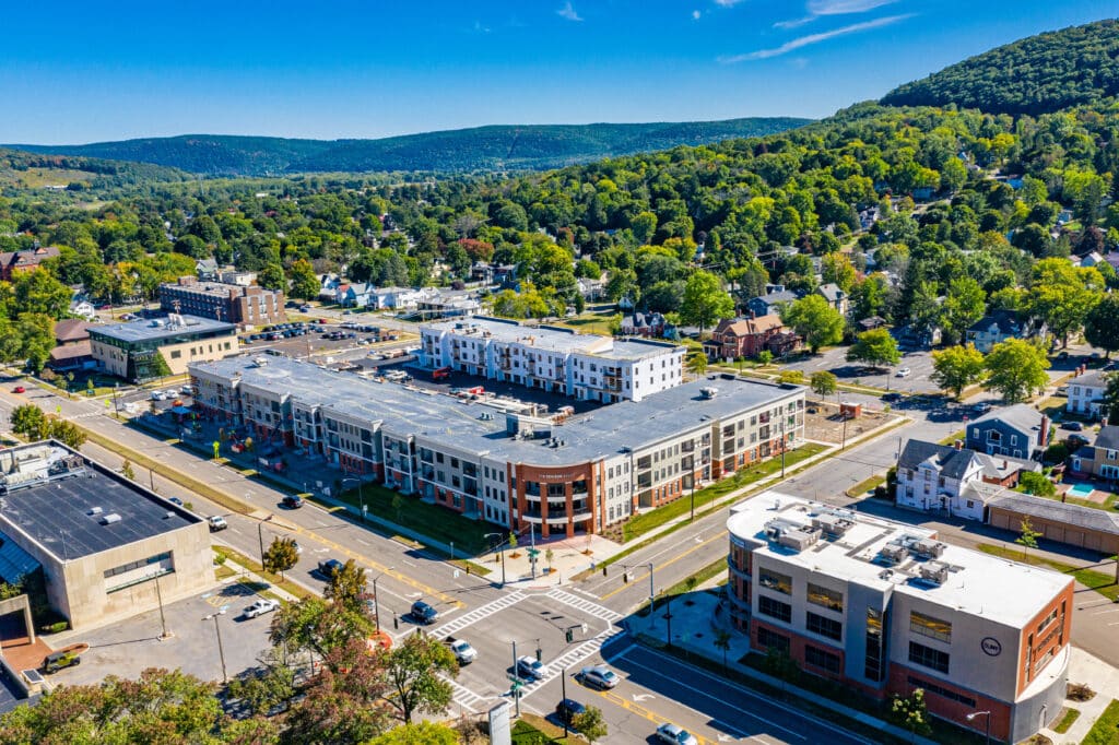 Aerial view of a small town with residential and commercial buildings, streets, parked cars, and a backdrop of rolling hills and green trees under a clear blue sky.