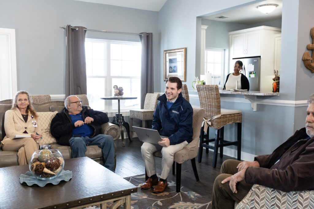 A group of people sitting and standing in a living room, one person with a laptop, and another standing in the kitchen area.