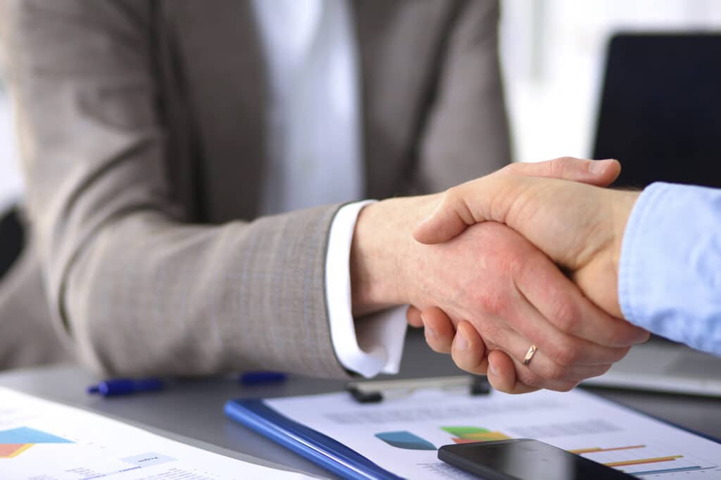 Two people shaking hands over a desk with documents, a pen, and a smartphone.