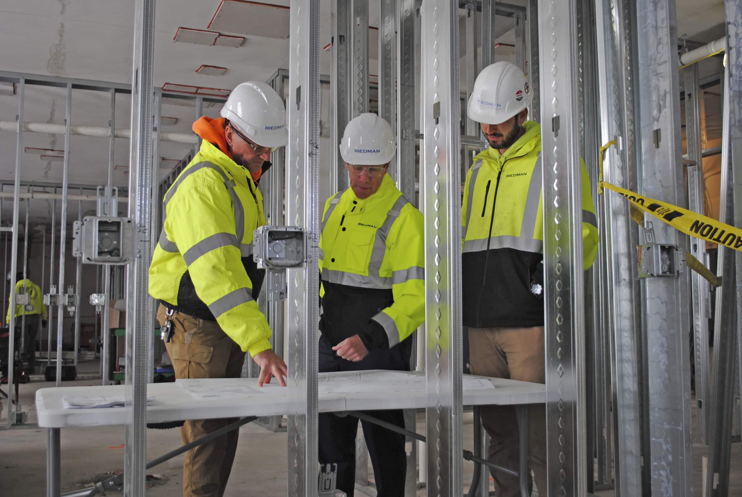 Three construction workers in safety gear review blueprints at a table inside a building under construction.