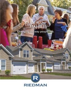 A group of people socializing outdoors in front of a house with a "Fairlawn Hills" sign and the text "Changing the way people live" above homes and a Riedman logo.