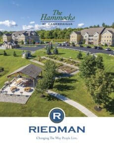 Aerial view of The Hammocks at Canandaigua, featuring apartments, a pavilion, and landscaping. Riedman logo and slogan "Changing The Way People Live" at the bottom.