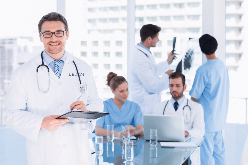 A group of medical professionals in a bright office. One doctor at the forefront holds a clipboard, while others discuss and review medical information on a laptop and X-ray.