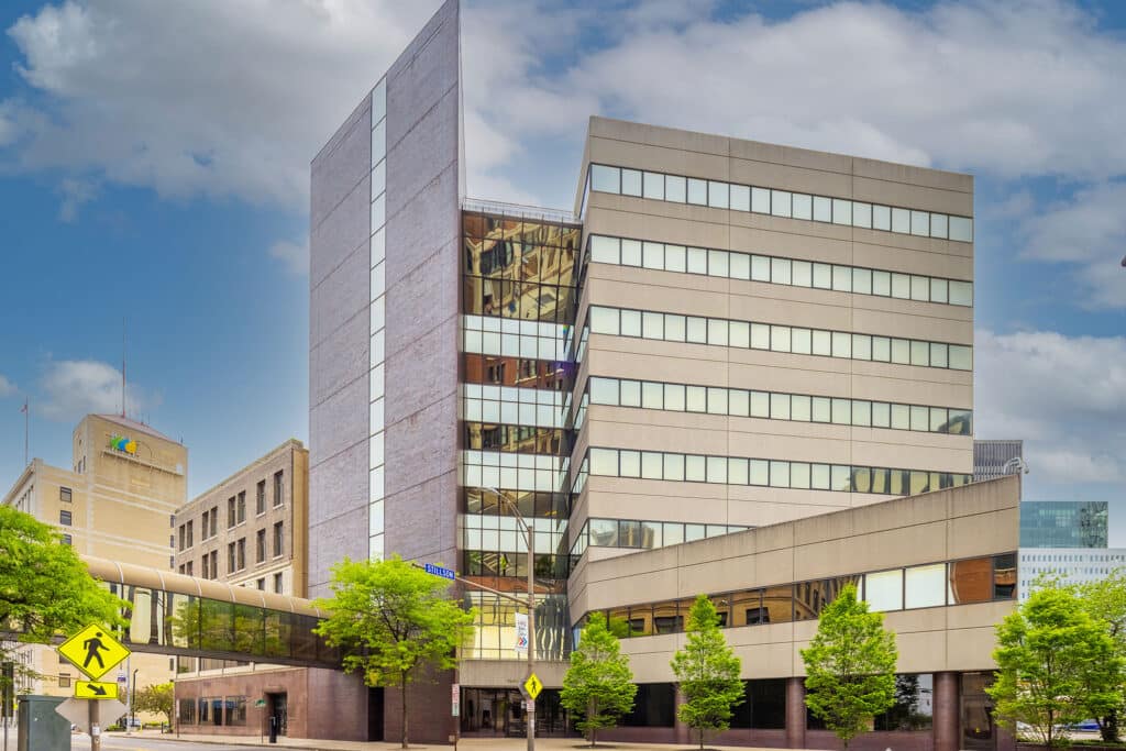 Modern office building with a skybridge connecting to another structure. The facade features glass and cement panels, with trees and a pedestrian crossing sign in the foreground.