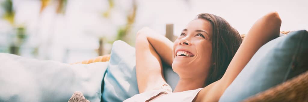 Smiling woman relaxing outdoors on a cushioned seat with arms behind her head.