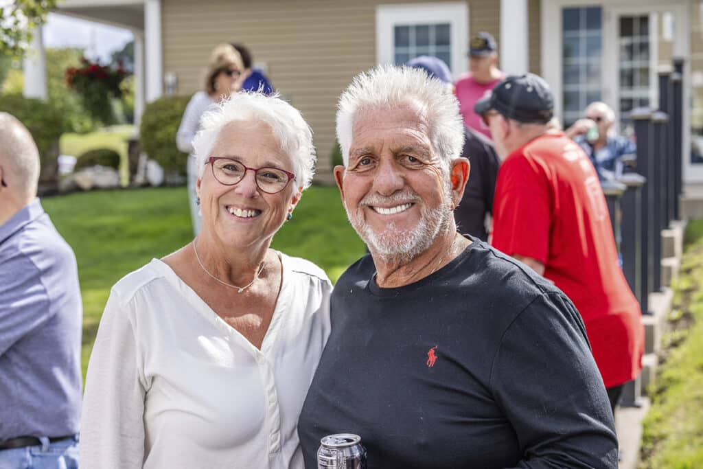 An older man and woman with white hair smile at the camera outdoors, with people blurred in the background.