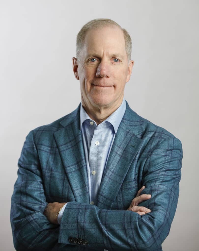 A middle-aged man with short light hair, wearing a blue plaid suit jacket and light shirt, stands with arms crossed against a plain light background.