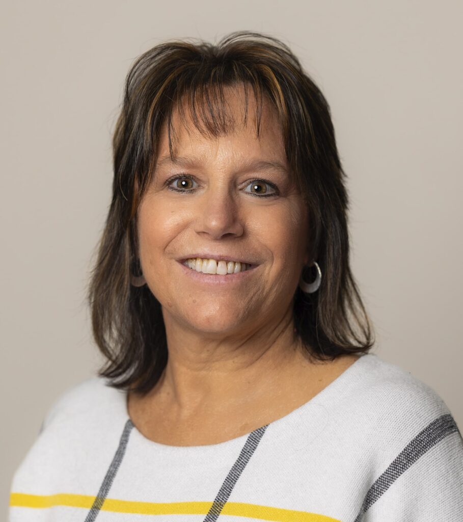 A woman with shoulder-length brown hair, wearing a white sweater with yellow and black stripes, smiles at the camera against a plain background.