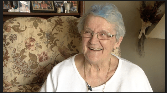 An elderly woman with glasses and short gray hair sits on a floral armchair, smiling. Framed photos and a floral arrangement are visible in the background.