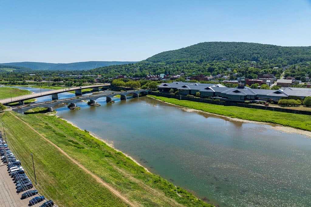 Aerial view of a river with a bridge, grassy banks, a parking lot, and a town with hills in the background under a clear blue sky.