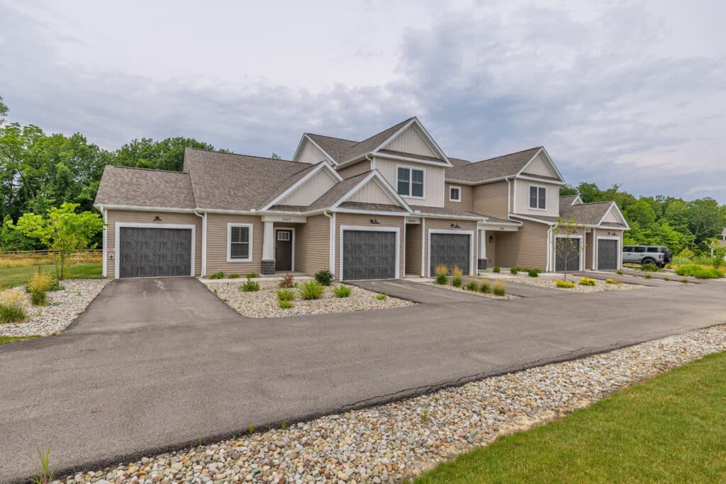 Modern townhouse view with attached garages, stone landscaping, and paved driveway under a cloudy sky, surrounded by greenery. Featuring private entrances.