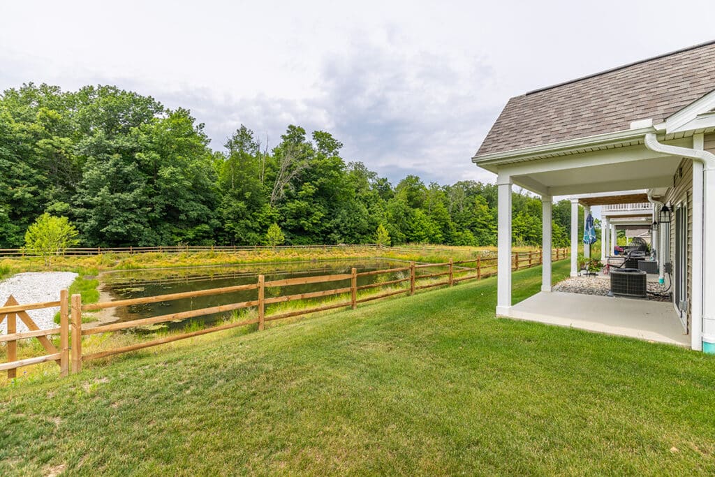 Covered patio overlooking a grassy yard, wooden fence, and a pond with trees in the background.