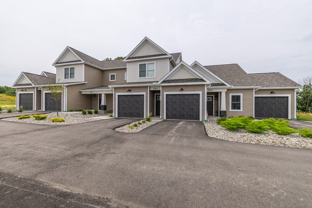 Two-story townhouse units with attached garages, grey siding, and landscaped front yards, set along a paved driveway.