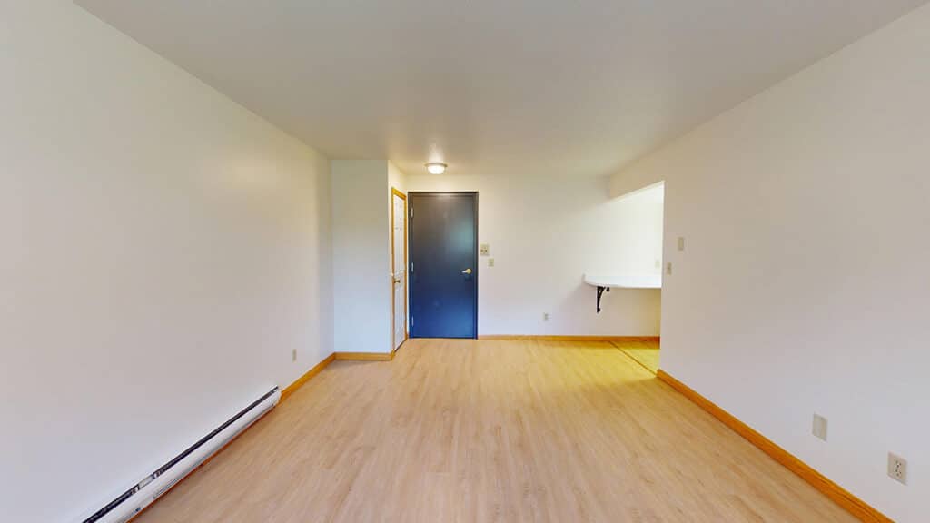 Living room and entryway inside a 1-bedroom home at Royal Villa Apartments in Erie, PA featuring wood-style flooring, baseboard heating, and neutral walls. The space includes a dark entry door, overhead lighting, and a pass-through opening to the kitchen, creating a functional and welcoming layout.