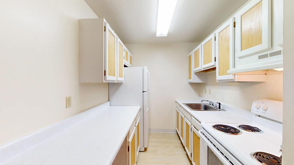 Galley kitchen at Royal Villa Apartments in Erie, PA featuring two-tone cabinets with natural wood accents, white appliances including refrigerator and electric stove, stainless steel sink, and bright overhead lighting. Spacious countertops provide ample room for meal prep in this rental home.