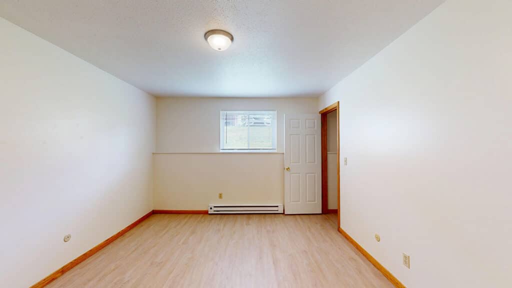 Inside a 1-bedroom apartment at Royal Villa Apartments in Erie, PA featuring LVP-style flooring, neutral walls, baseboard heating, and a bright window with vertical blinds. The open layout offers a versatile space for comfortable apartment living.