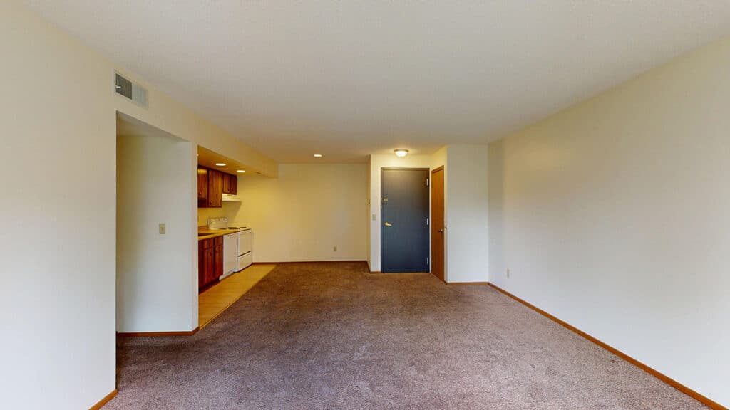 Open-concept living area at Royal Villa Apartments in Erie, PA featuring plush carpeting, neutral walls, and recessed lighting. The space connects seamlessly to the kitchen with wood cabinetry, white appliances, and an entryway with closet storage, creating a functional apartment layout.