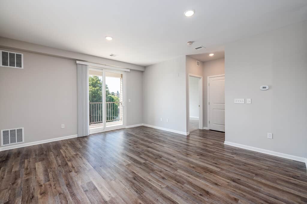 Unfurnished living room with wood flooring, light gray walls, recessed lighting, sliding glass door to balcony, and a door leading to the bedroom.