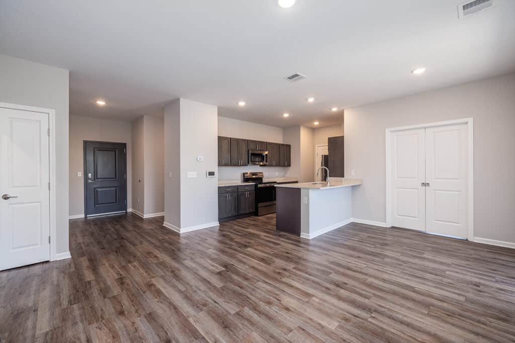 Modern, unfurnished apartment with wood-look LVP flooring, white walls, open kitchen with dark cabinets, quartz countertops, stainless steel appliances with a breakfast bar.