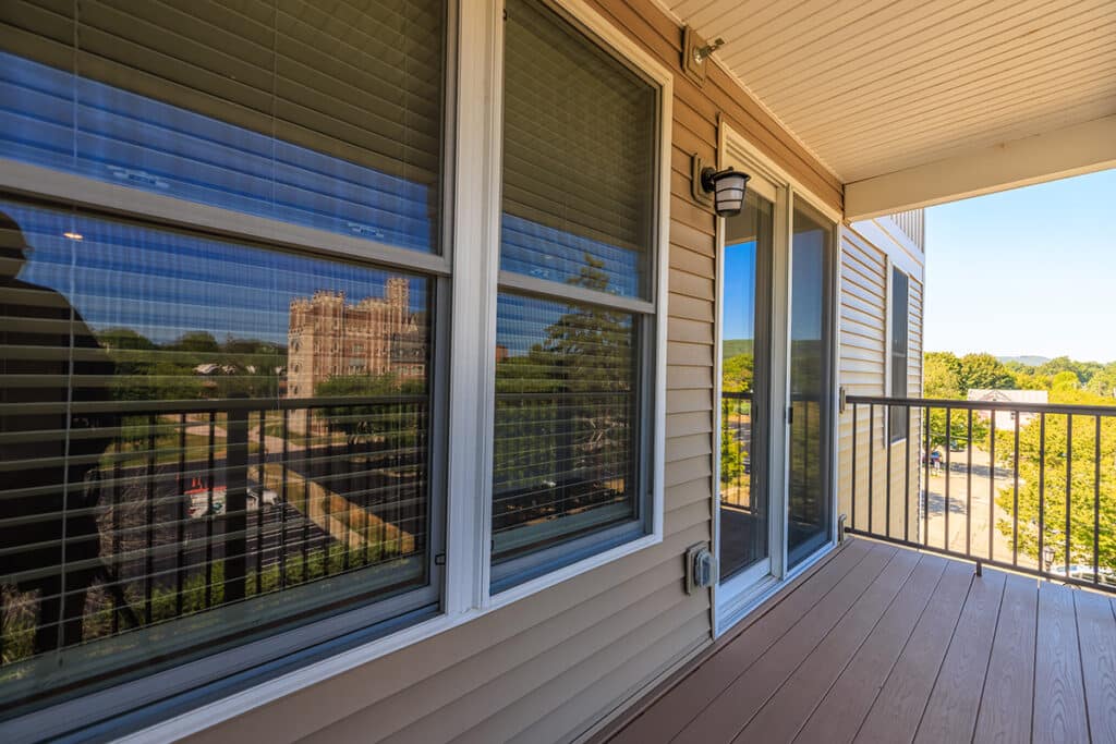 A covered balcony with trex flooring, beige siding, large windows with blinds, and a glass sliding door overlooking a green, overlooking the natural landscape of Elmira NY.
