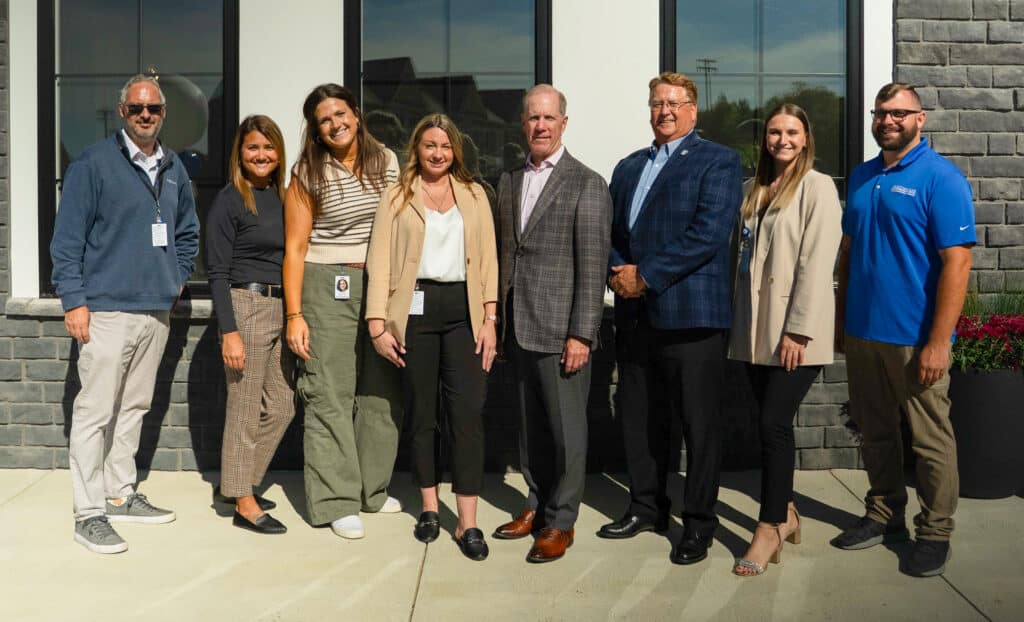 A group of eight people, four men and four women, stand side by side outdoors in front of a building, posing for a photo in business-casual attire.