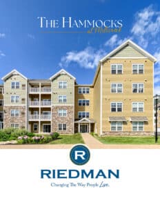 A modern, four-story apartment building with balconies and stone accents, under a blue sky, with the text "The Hammocks at Millcreek" and "Riedman" below.