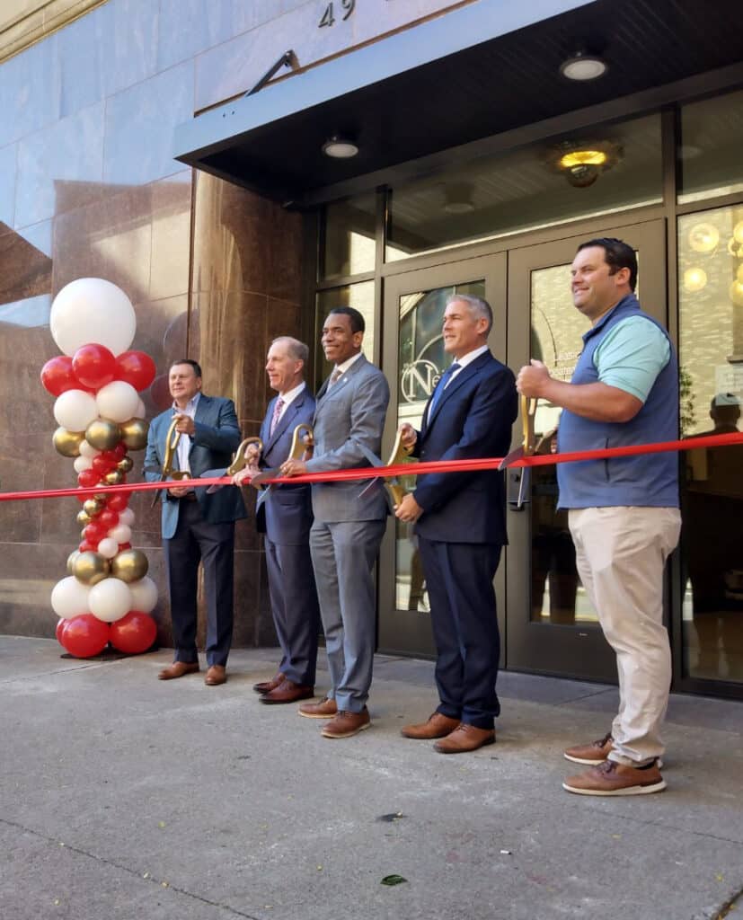 Five men in business attire stand in front of The Neisner at East End, holding large scissors and cutting a red ribbon at a ribbon-cutting ceremony. Balloon columns are visible on the left at Rochester Apartments.