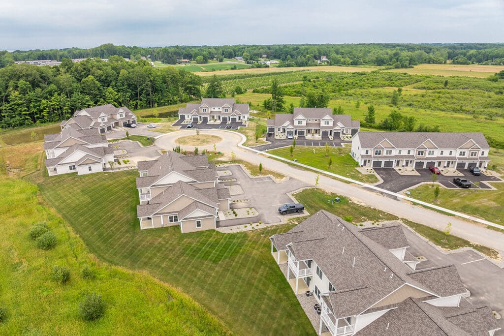 Aerial view of a suburban housing complex with multiple buildings, driveways, and green lawns surrounded by fields and trees under a cloudy sky.