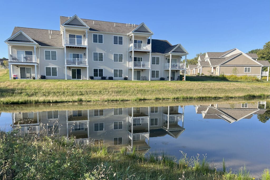 Three-story apartment buildings with balconies reflecting on the pond at The Hammocks at Fairview, surrounded by green space and serene wooded scenery.