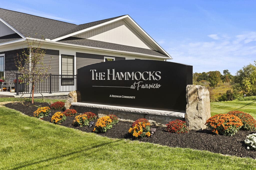 Front entrance sign for The Hammocks at Fairview, a Riedman community, featuring a modern black monument surrounded by colorful landscaping and located beside the resident clubhouse.