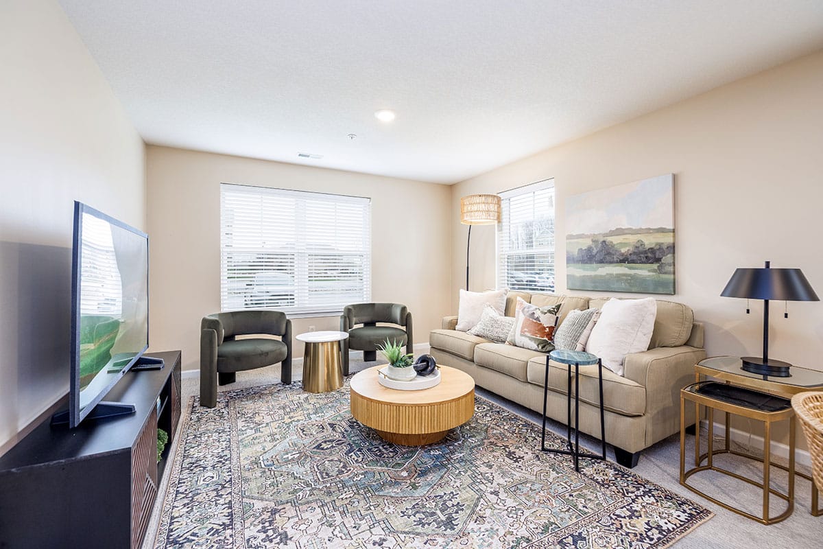 Bright living room with a beige sofa, two dark armchairs, a large patterned rug, a round coffee table, TV, and wall art, lit by natural light from two windows.