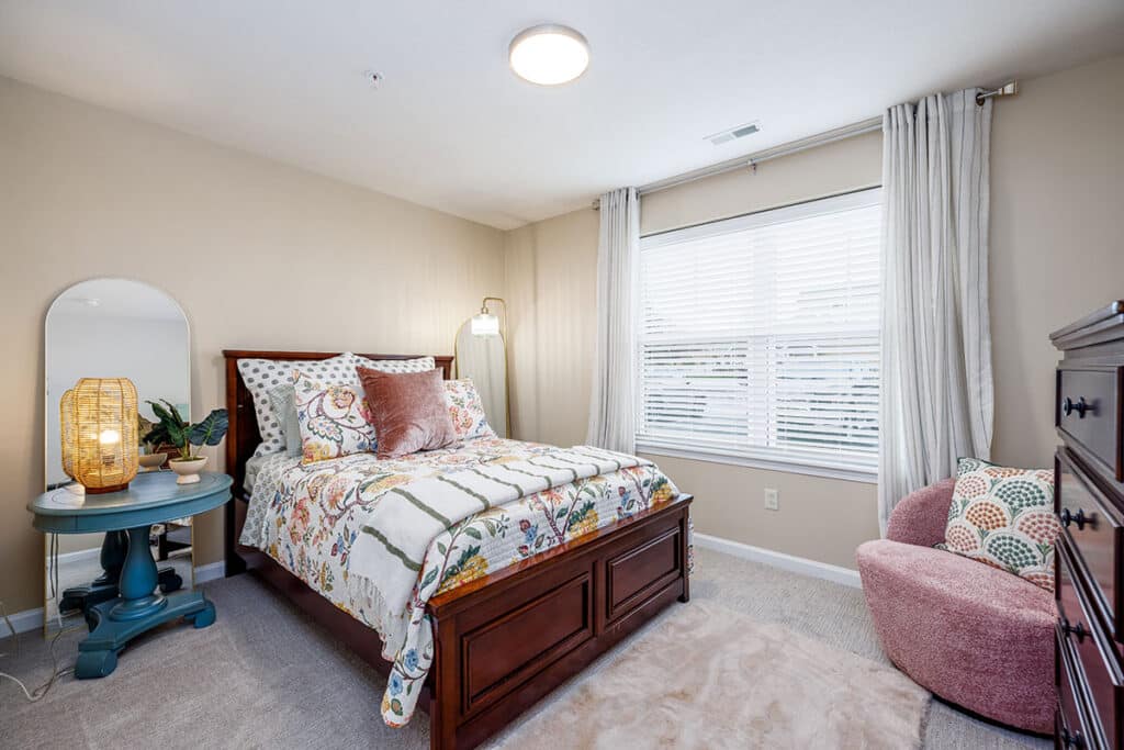 Guest bedroom with dark wood bed, patterned bedding, large window, neutral walls, and accent chair at The Hammocks at Millcreek