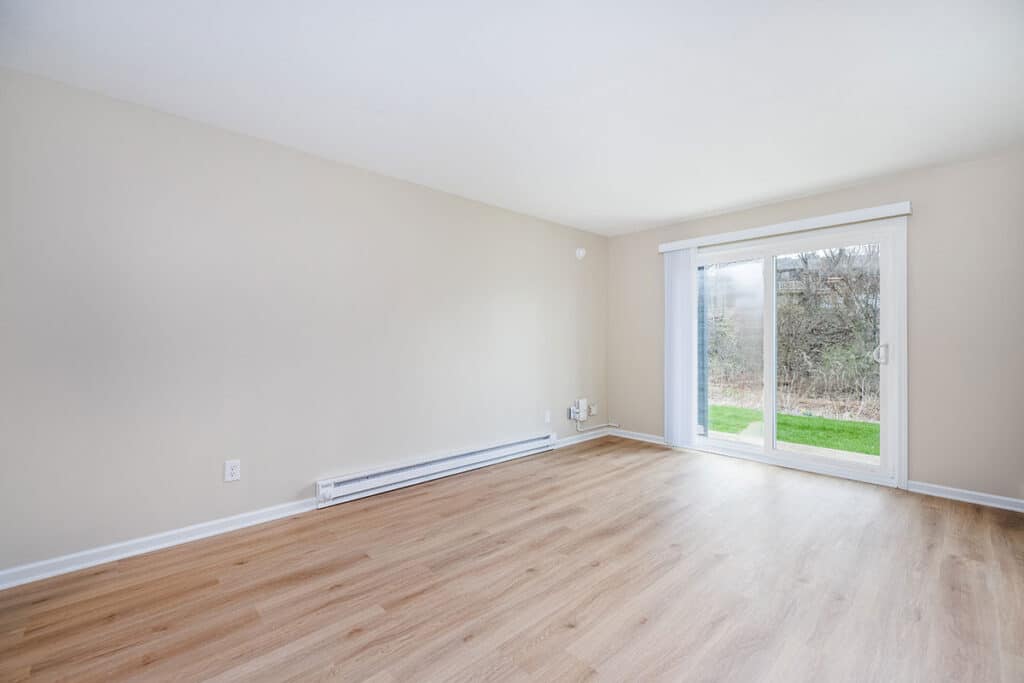 Bright unfurnished living room at Royal Villa Apartments in Erie, PA with wood-style flooring and sliding glass doors to outdoor green space.